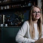 A smiling young woman with eyeglasses sitting in a stylish coffee shop interior.