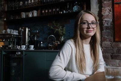 A smiling young woman with eyeglasses sitting in a stylish coffee shop interior.