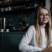 A smiling young woman with eyeglasses sitting in a stylish coffee shop interior.
